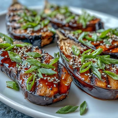 Two roasted Japanese eggplant halves with a glossy, deep amber miso glaze and toasted sesame seeds, served warm on a white plate.  
